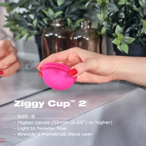 Rosa Menstruationscup wird von einer Person gehalten, im Hintergrund grüne Pflanzen und braune Flaschen.