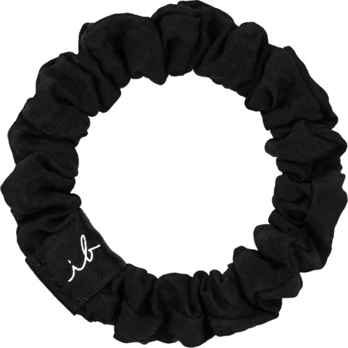 A black fabric hair tie with a small white signature or logo on it. It is circular and scrunches to hold hair.