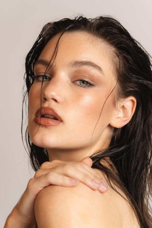 A woman with wet hair gazes softly, hand on shoulder, makeup emphasizes her face, neutral background.