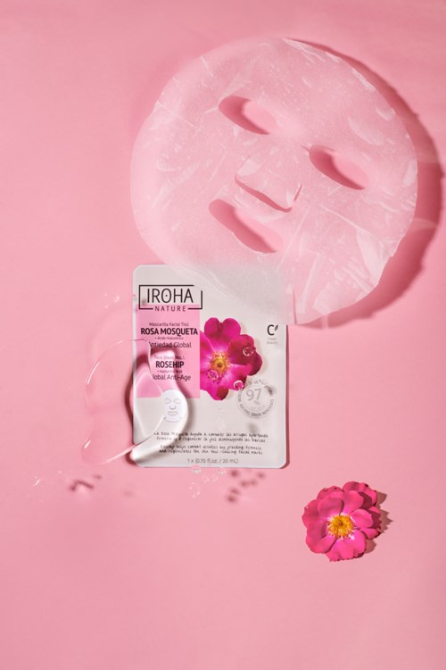 Een roze gezichtsmasker met oog- en mondgaten, een stuk zeep en een roze bloem op roze achtergrond.