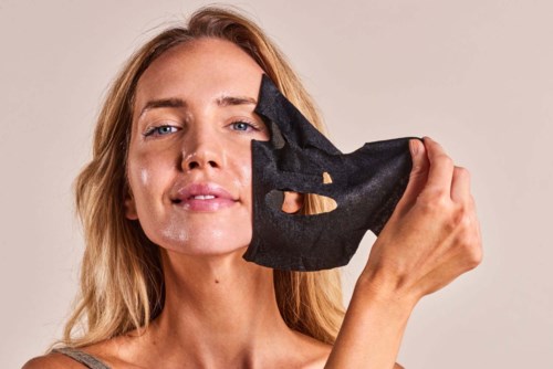 Een vrouw houdt een zwart stoffenmasker dicht bij haar gezicht. Ze heeft licht haar en een zelfverzekerde uitdrukking.