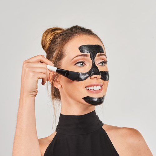 Een vrouw met een zwarte gezichtsmasker, houdt een kwast dichtbij haar wang, glimlacht. Ze draagt een zwart topje.