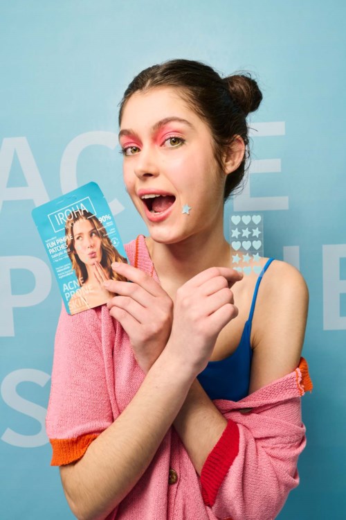 Een jonge vrouw met opvallende make-up houdt een testkaart vast, glimlacht en draagt kleurrijke kleren voor een blauwe achtergrond.