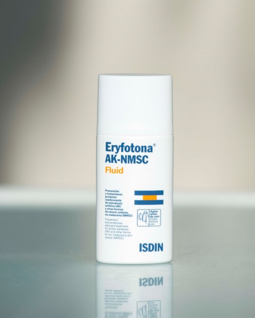 Eine weiße Flasche mit blauer und orangener Beschriftung, gekennzeichnet als Eryfotona AK-NMSC Fluid von ISDIN, steht auf einer reflektierenden Oberfläche.