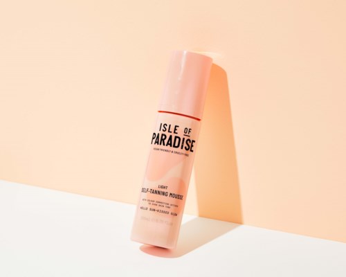 A pink bottle of Isle of Paradise self-tanning mousse with a simple design, leaning against a peach-colored wall.