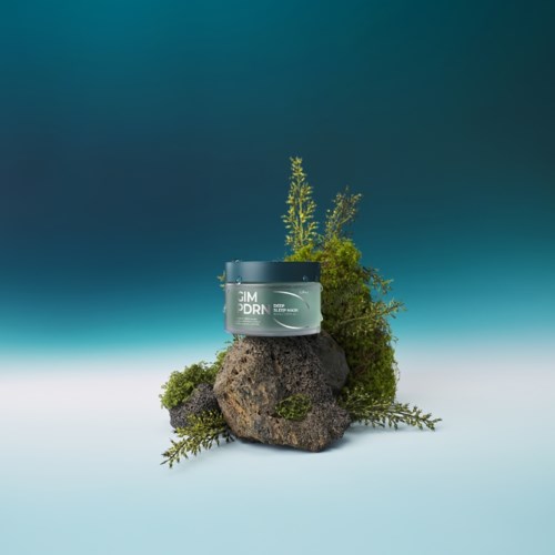 Een zwarte container met "Skim Dren" erop, op stenen met groene planten en een blauwgroene achtergrond.