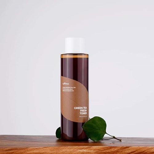 Een lange bruine fles met label 'Green Tea Fresh Toner' staat op een houten oppervlak met groene bladeren ernaast.