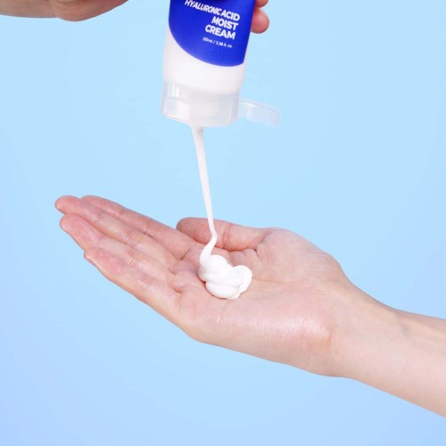 A hand holds a small amount of cream from a bottle labeled 'Hyaluronic Acid Moist Cream' against a blue background.
