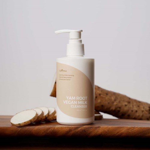 Eine weiße Flasche mit Pumpkopf, beschriftet mit 'YAM ROOT VEGAN MILK CLEANSER' auf einer Holzfläche, mit Yam-Scheiben und Yamwurzel daneben.