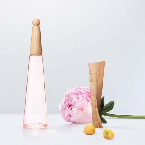 Een roze parfumfles, een roze pioenroos, een houten object en kleine gele en roze vruchten op een witte achtergrond.