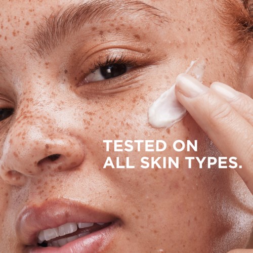 Close-up of a woman with freckles applying a cream near her eye. Text states the product is tested on all skin types.