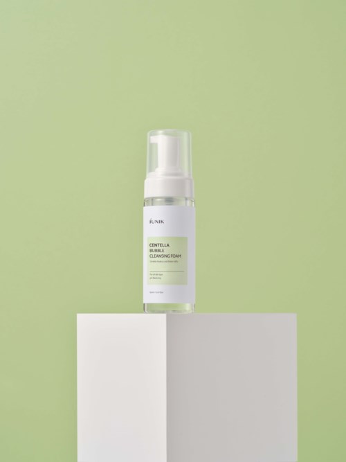 Een doorzichtige fles met een witte label en pomp, op een witte blok tegen een groene achtergrond.