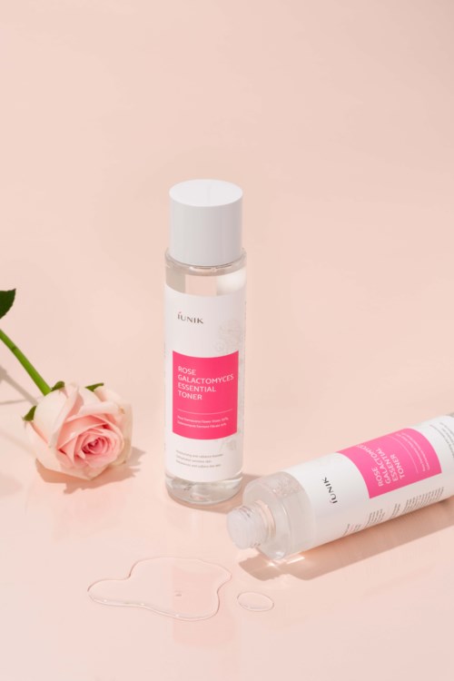Twee flessen gezichtstoner met roze labels, één ligt en één staat naast een roze roos.