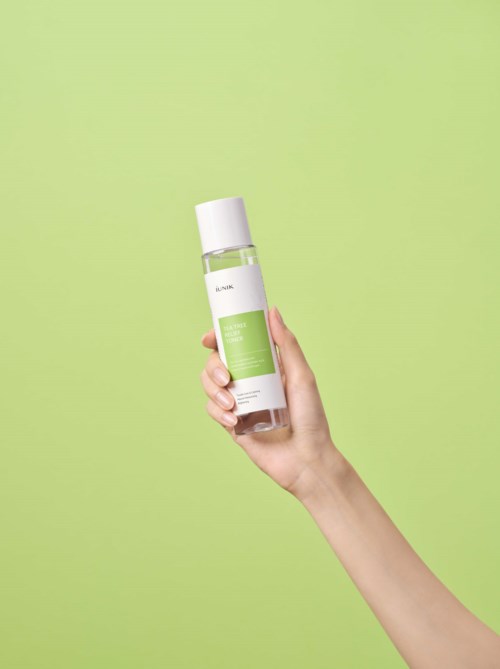 Een hand houdt een doorzichtige fles met een witte dop, groen label en heldere vloeistof, tegen een lichtgroene achtergrond.