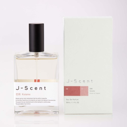 En genomskinlig parfymyssha med svart kork och J-scent-logga bredvid sin vita box med minimalistisk design och ljusrosa detaljer.