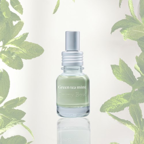 Een kleine glazen fles met het label Green Tea Mint omgeven door muntbladeren, met een eenvoudige lichte achtergrond.