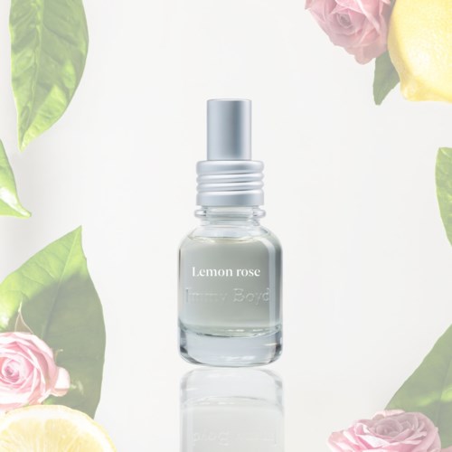 Klein glazen flesje met label Lemon Rose en spraydop, omgeven door roze rozen en citroenpartjes.