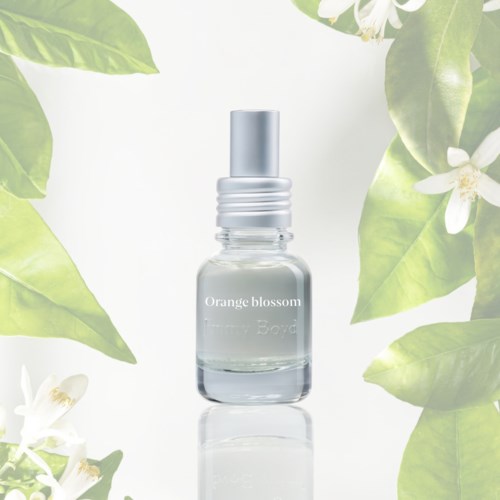 Een klein heldere sprayfles met label 'Orange blossom' omgeven door groene bladeren en witte bloemen.