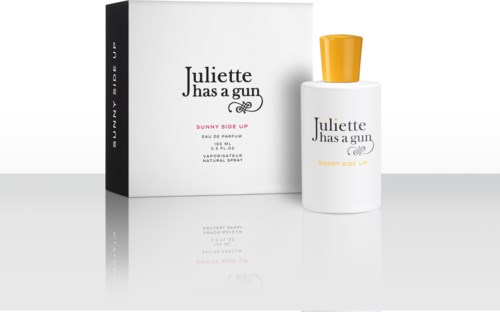 Een witte fles parfum met een gele dop naast een bijpassend doosje. De fles heeft zwarte en gele tekst.