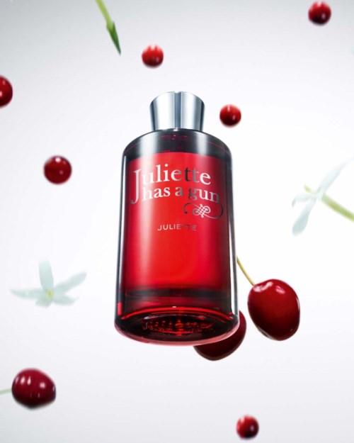 Eine rote Parfümflasche mit Kirschen und grünen Blättern darin, die in der Luft schwebt. Die Flasche trägt die Aufschrift "Juliette Has a Gun."