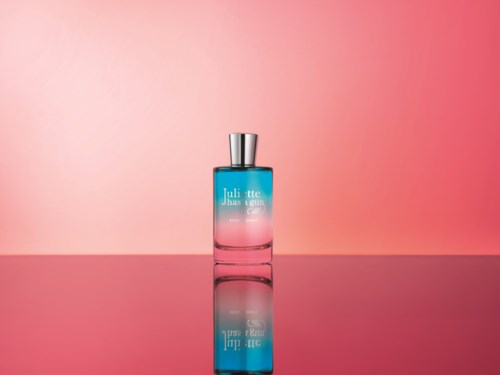 A blue and pink gradient perfume bottle with a silver cap, placed on a reflective surface against a pink background.