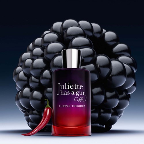 A perfume bottle labeled Juliette Has A Gun with purple and red gradient, placed in front of a bunch of black grapes and a red chili pepper.