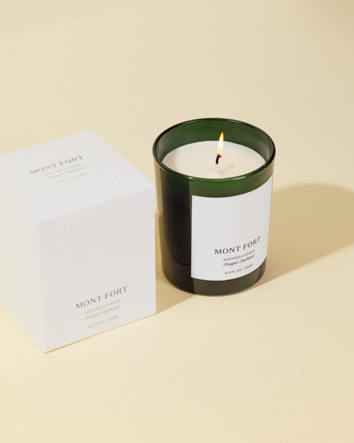 A lit candle with a black glass holder next to a white box, both labeled Mont Fort, on a light background.