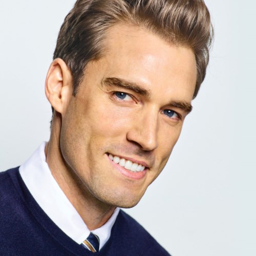 A smiling man with blue eyes and brown hair, wearing a navy sweater and white shirt.