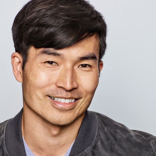 A smiling man with black hair wearing a dark jacket and a light shirt in front of a plain background.