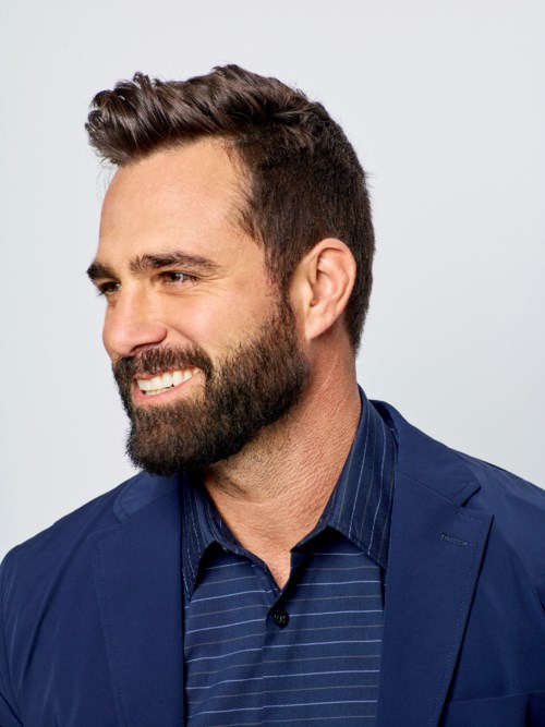 A smiling man with a beard in a blue jacket and striped shirt against a light background.