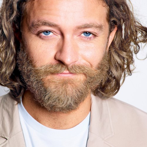 A man with long, curly hair, a full beard, and blue eyes looks directly at the camera.