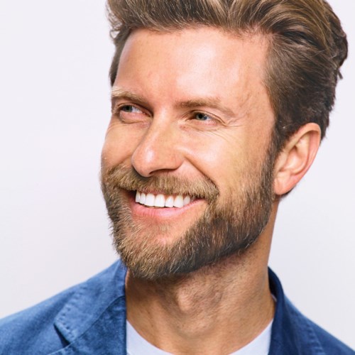 A smiling man with a beard and light brown hair, wearing a blue blazer, looks to the side.