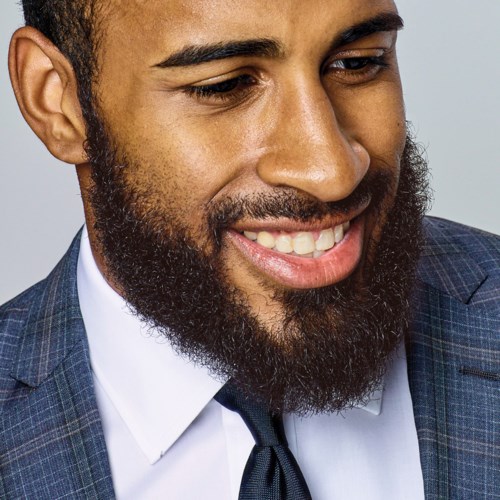A man with a beard smiling, wearing a suit and tie, looking to the side.