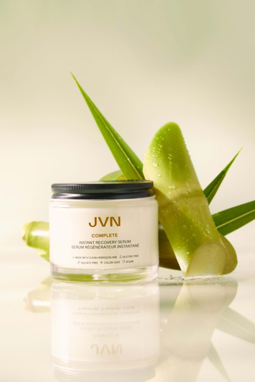 Ein Glas JVN Complete Serum mit Aloe Vera Blättern und Scheiben im Hintergrund.