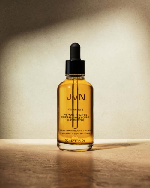 Kleine bernsteinfarbige Glasflasche mit Pipettenverschluss, beschriftet 'JMN Complete Pre-Wash Scalp Oil,' steht auf einer Holzoberfläche.