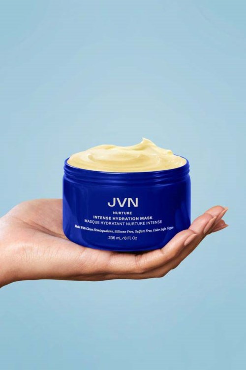 Eine Hand hält einen blauen Behälter mit Haarmaske und Creme gegen hellblauen Hintergrund.