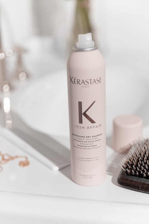 Vaaleanpunainen Kérastase-shampoopullo, harja ja peili kylpyhuoneen pöydällä.