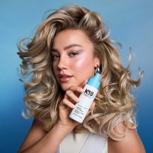 A woman with long, curly blonde hair holds a white skincare bottle in front of a blue background.
