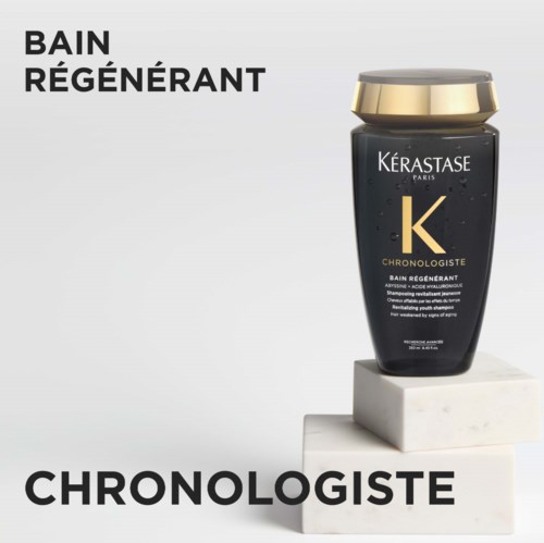 Een zwarte shampoefles met gouden dop van Kerastase Paris staat op een witte blok. Tekst bevat "Bain Regenerant" en "Chronologiste."