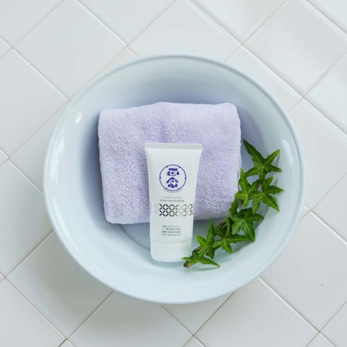 A white tube of skincare product resting on a neatly folded light purple towel in a white bowl, with ivy leaves beside.