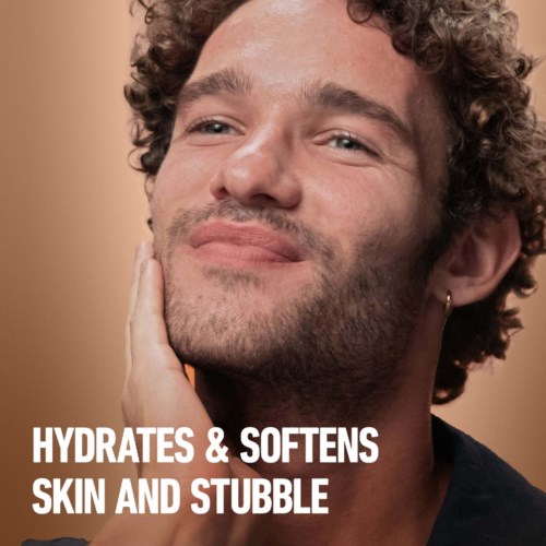 Ein Mann mit lockigem Haar, der sein Gesicht berührt, lächelt sanft. Text: "Hydrates & softens skin and stubble."