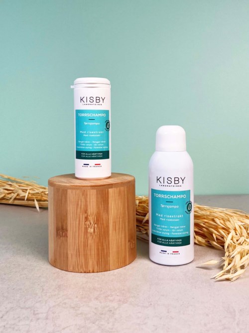 Two white bottles of KISBY shampoo on a wooden block, with dry wheat and a pastel background.