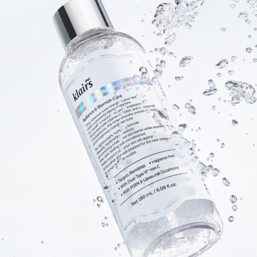 Een doorzichtige fles met pomp ligt op een witte ondergrond met waterdruppels eromheen. Het label toont huidverzorgingsinformatie.