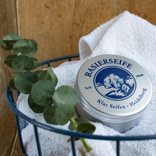 A round tin labeled "RASIERSIEFE" with a mountain scene, placed on a white towel inside a basket.