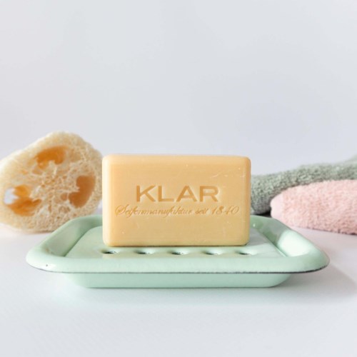 A bar of soap with the word "KLAR" engraved, placed on a soap dish with towels in the background.