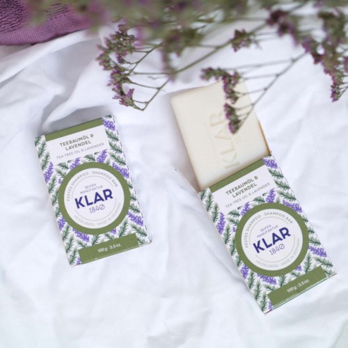 Bar soap with a white color, floral packaging, labeled 'Klar 1840', placed on white fabric with purple flowers.