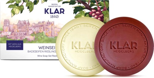 Two round soap bars, one white and one red, with 'KLAR' written on them, in front of a colorful box with grapes and a city.