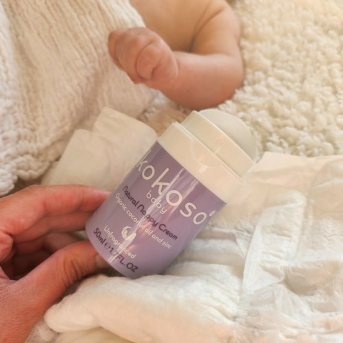 A person holds a purple bottle of baby cream next to a baby's hand and blanket.