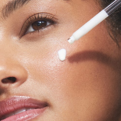 Eine Person trägt Hautpflege oder Make-up mit einer Pipette in der Nähe ihres Auges auf. Die Haut sieht klar und glatt aus.