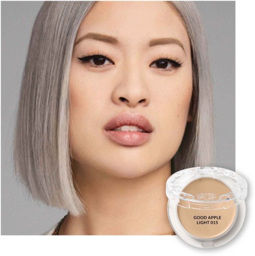 En kvinna med rakt grått hår och smink, visar ett neutralt uttryck. En liten burk av beige foundation finns längst ner till höger.